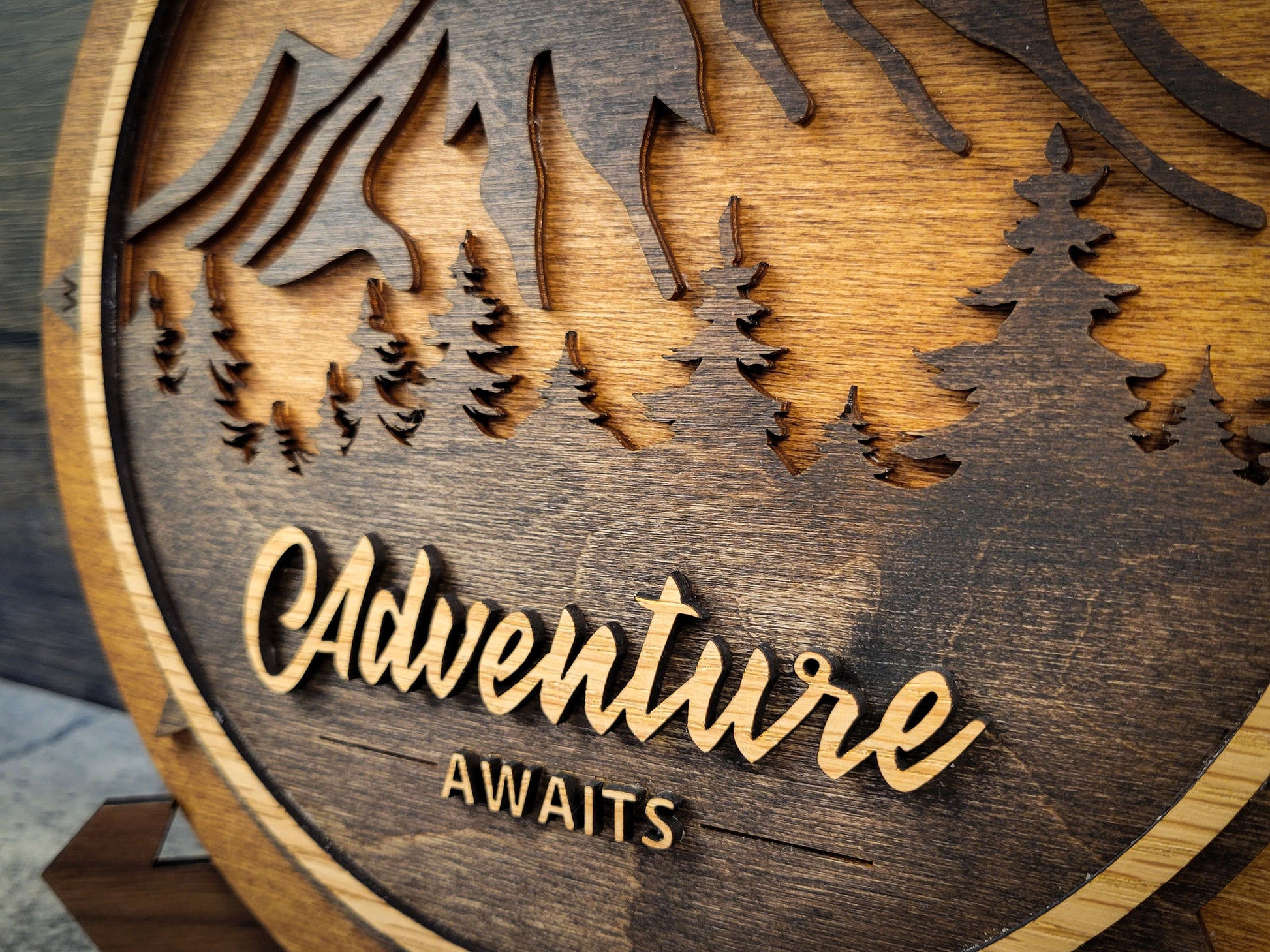 Wooden plaque with cut-out design of trees and mountains and the text 'Adventure Awaits'.