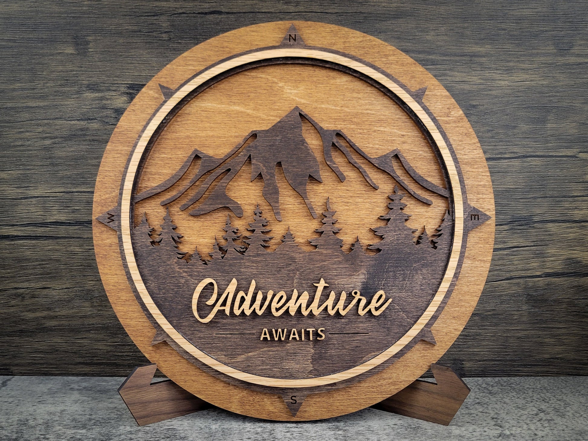 Wooden plaque with mountain design and 'Adventure Awaits' text on a wooden background