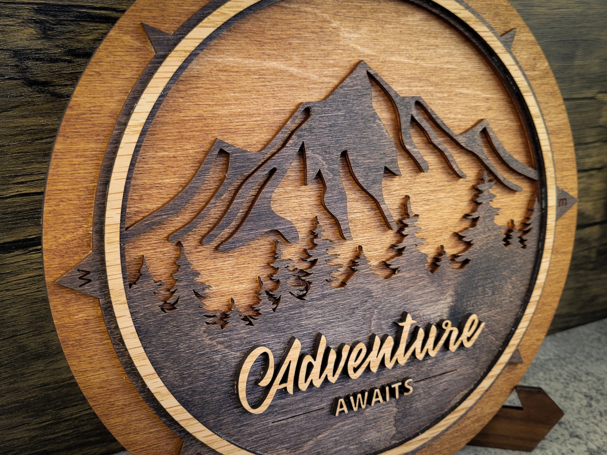 Wooden plaque with mountain design and 'Adventure Awaits' text on a wooden background