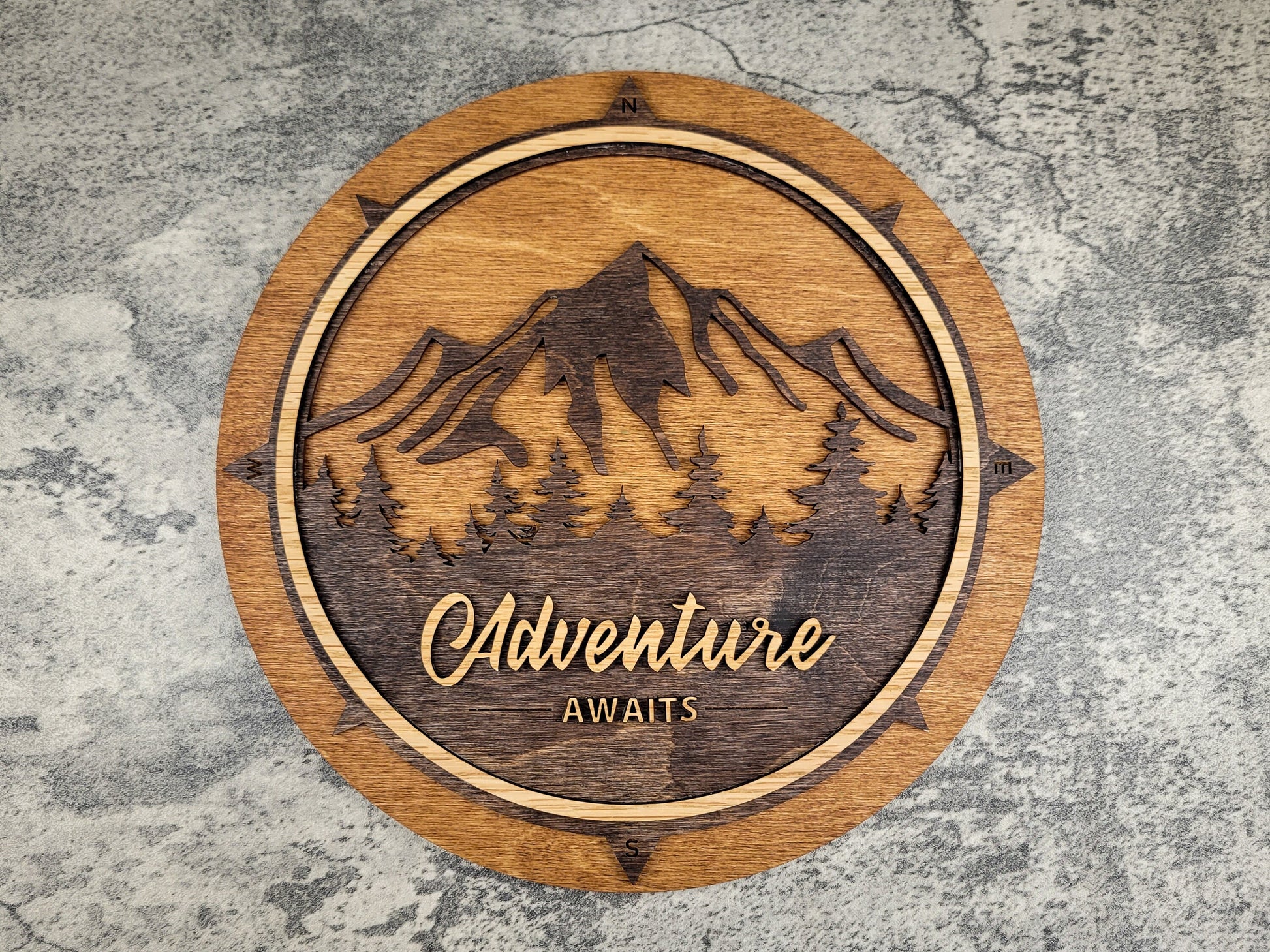 Wooden plaque with mountain design and 'Adventure Awaits' text on a textured surface