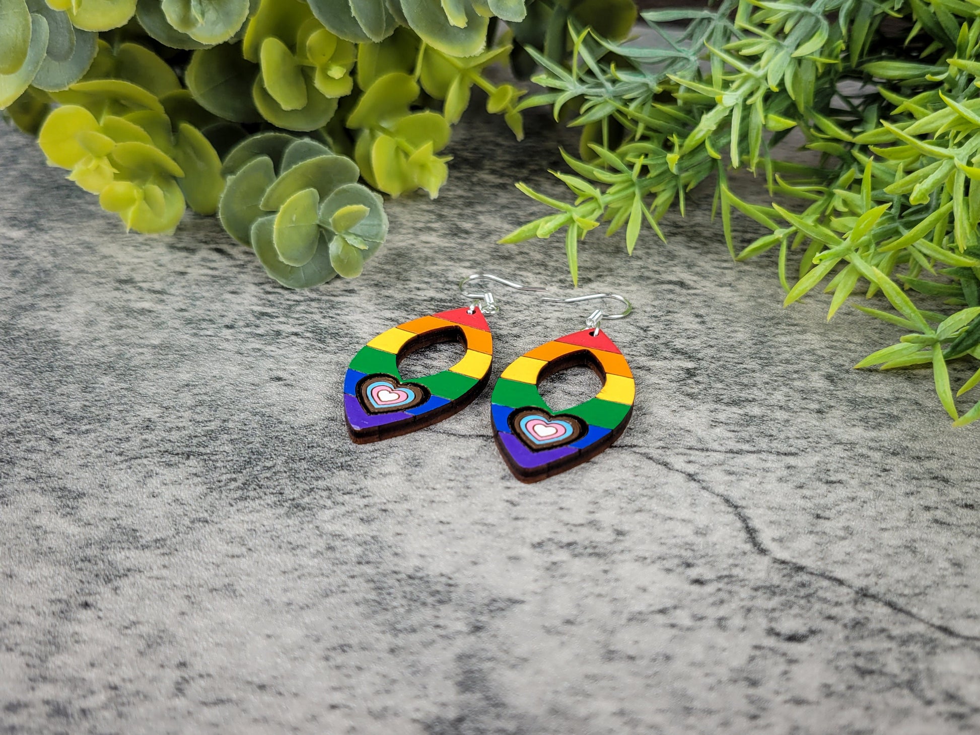 Rainbow heart-shaped earrings on a textured surface with green foliage in the background