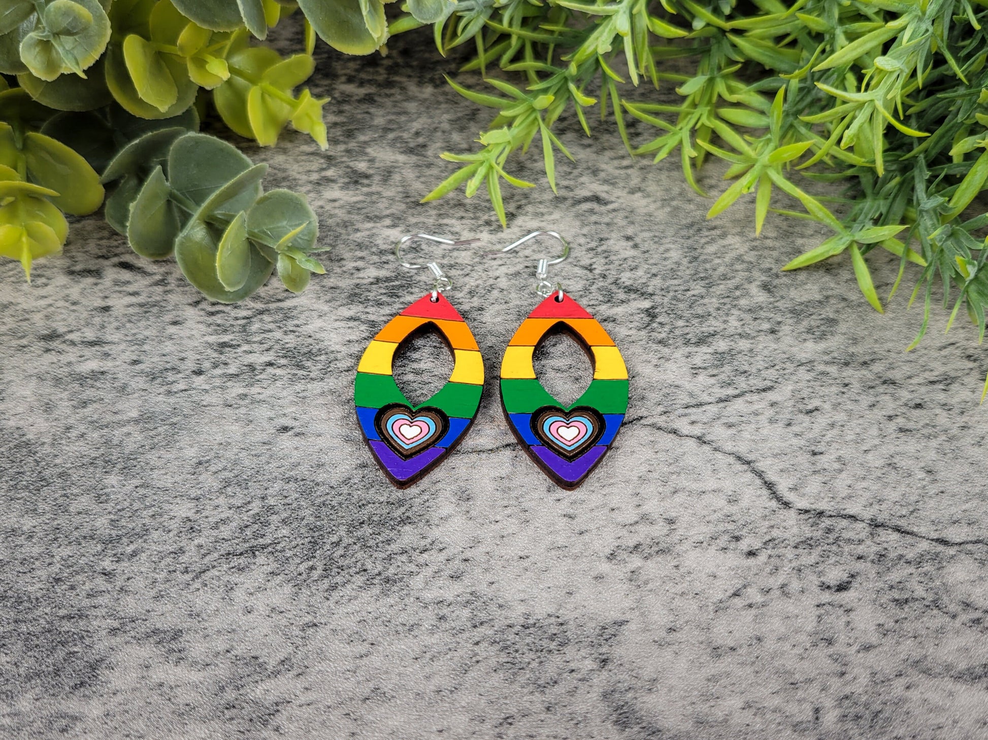 Rainbow heart-shaped earrings on a textured surface with greenery