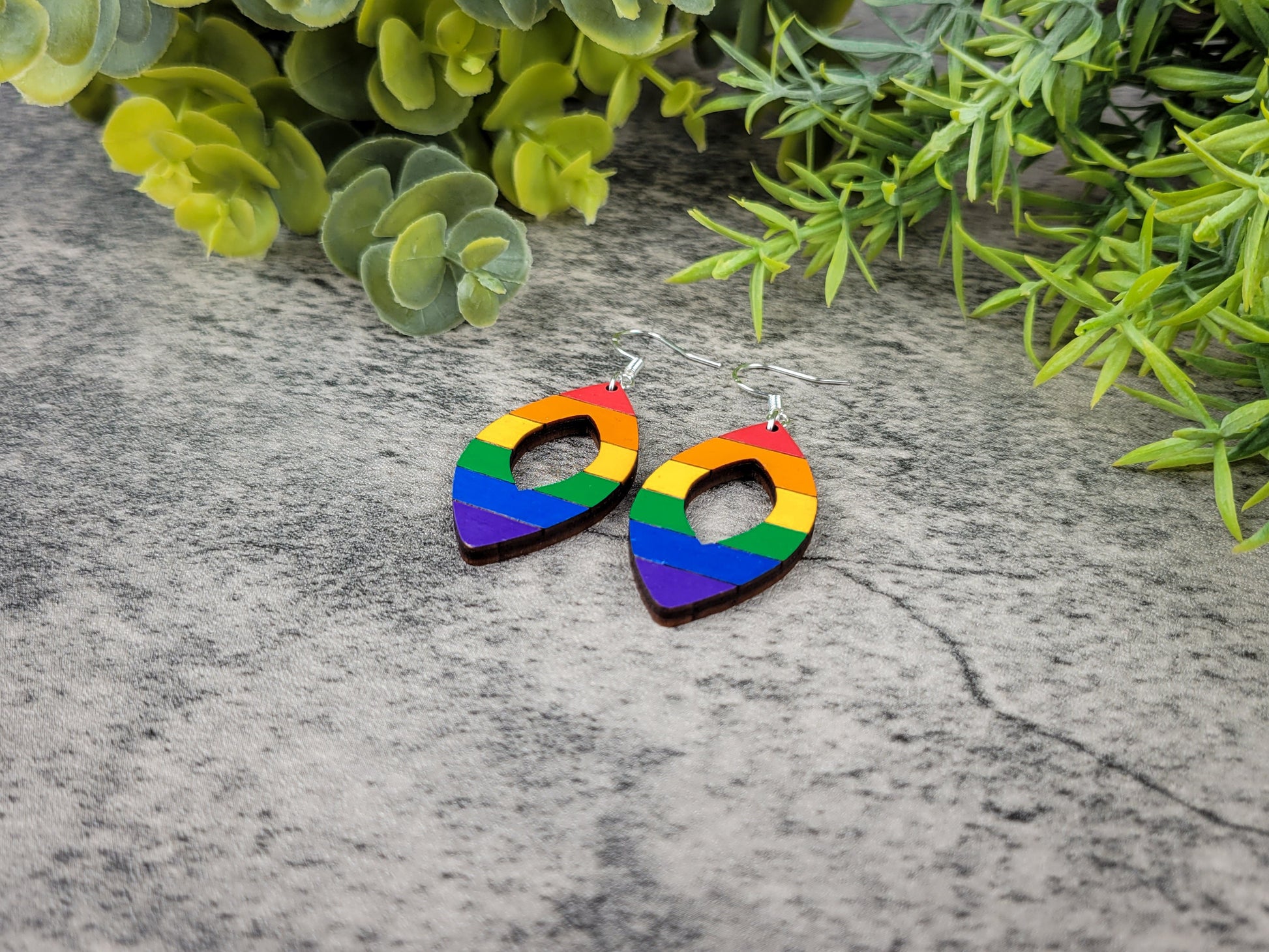 Rainbow heart-shaped earrings on a textured surface with green foliage in the background, angled view