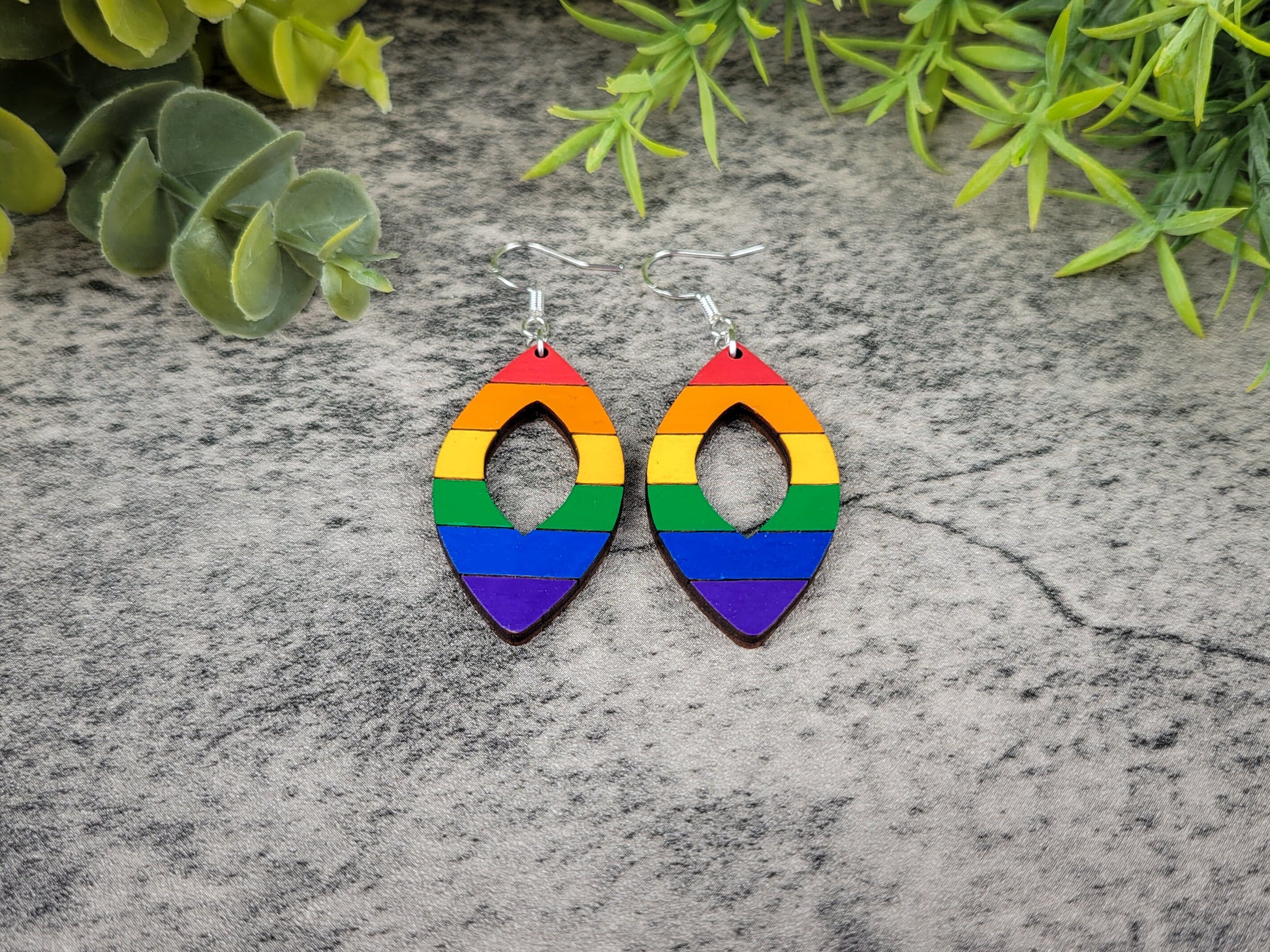 Rainbow-colored earrings on a textured stone surface with greenery in the background, front view