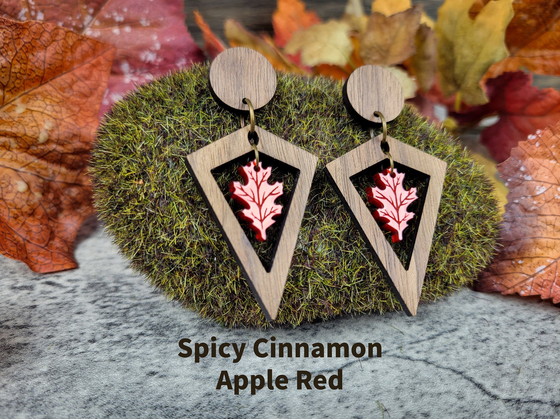 Wooden earrings with red leaf design on a moss ball with autumn leaves in the background