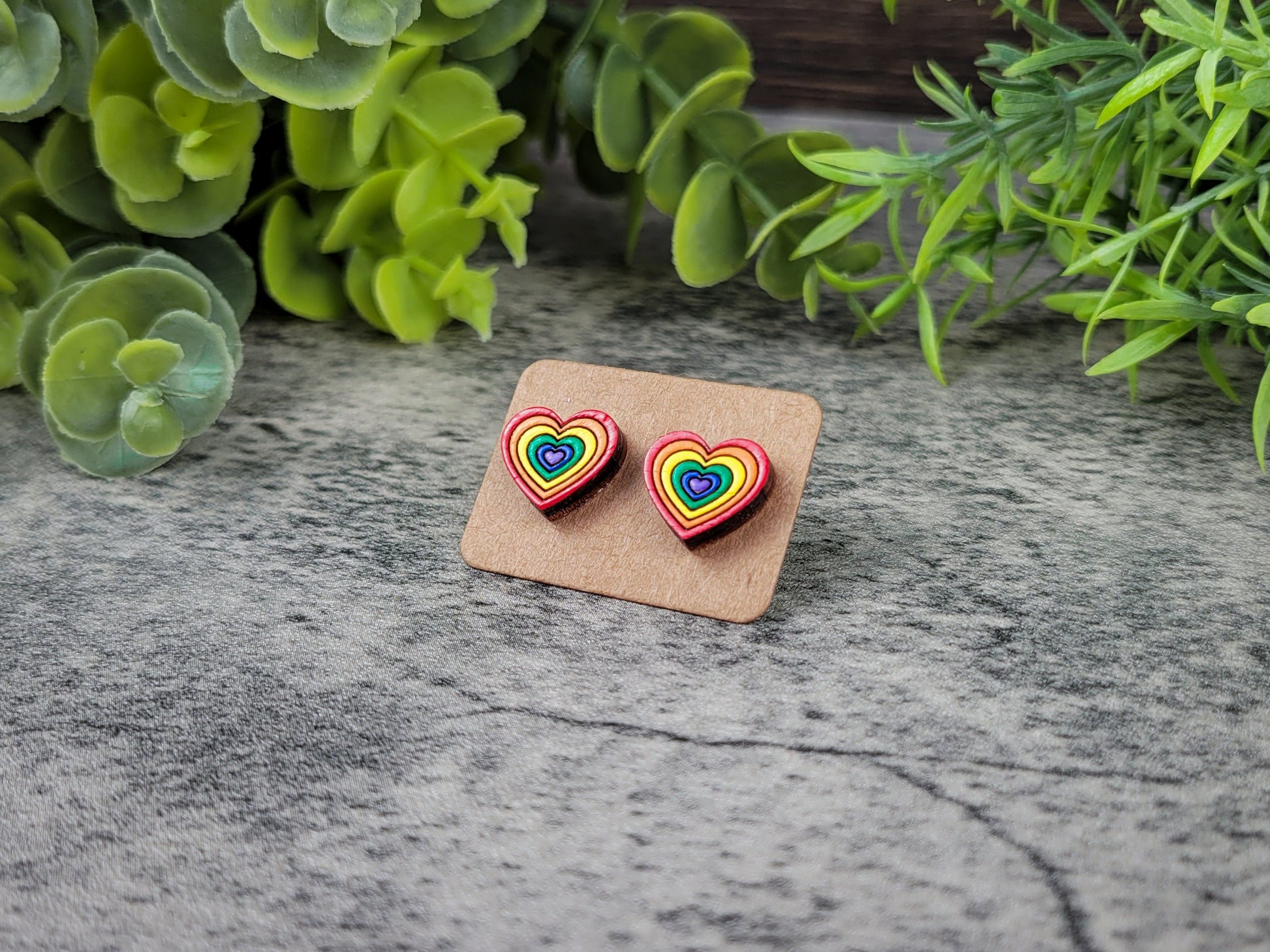 Rainbow heart-shaped earrings on a cardboard backing with green foliage in the background