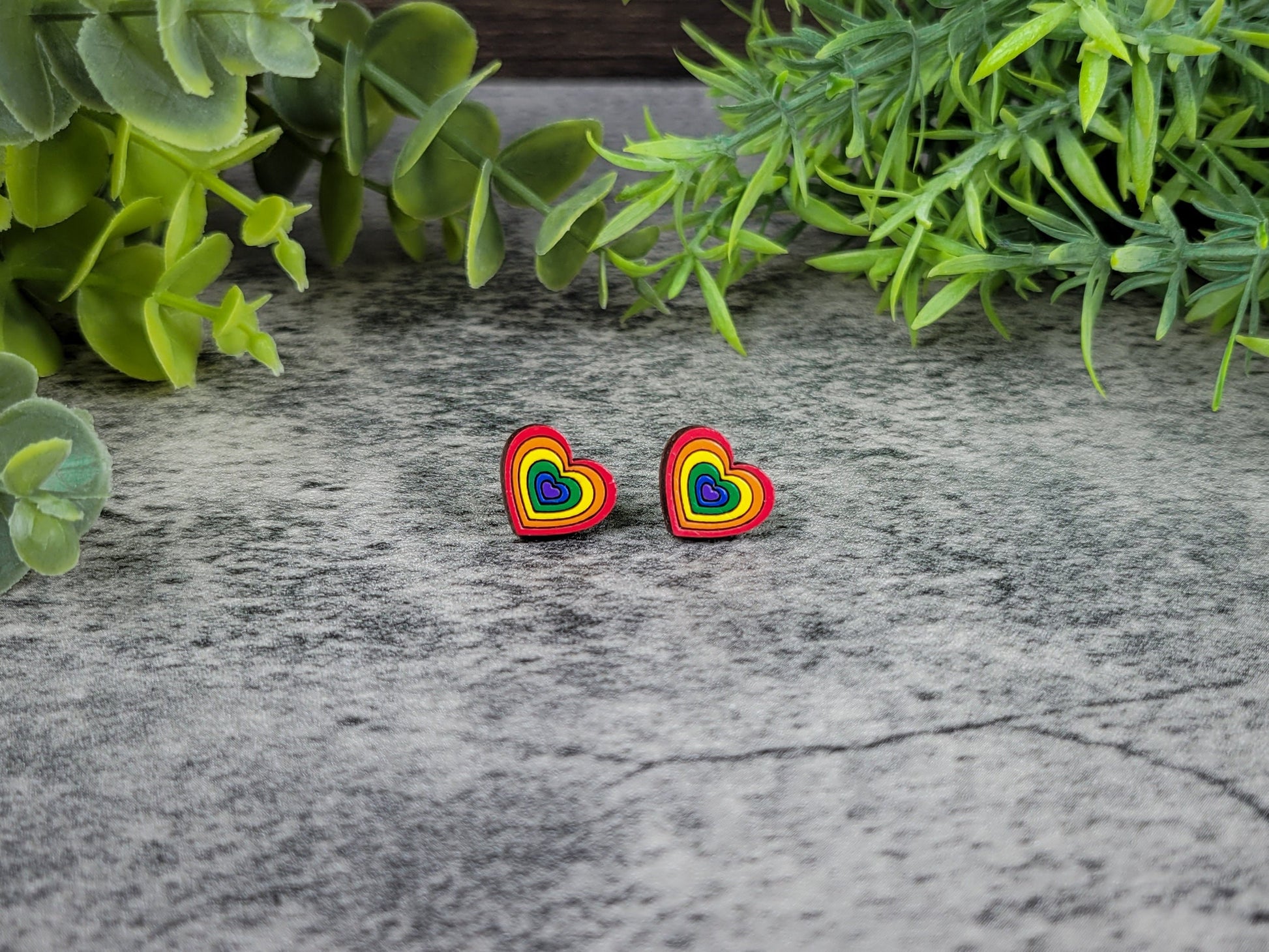 Rainbow heart-shaped earrings on a textured surface with green foliage