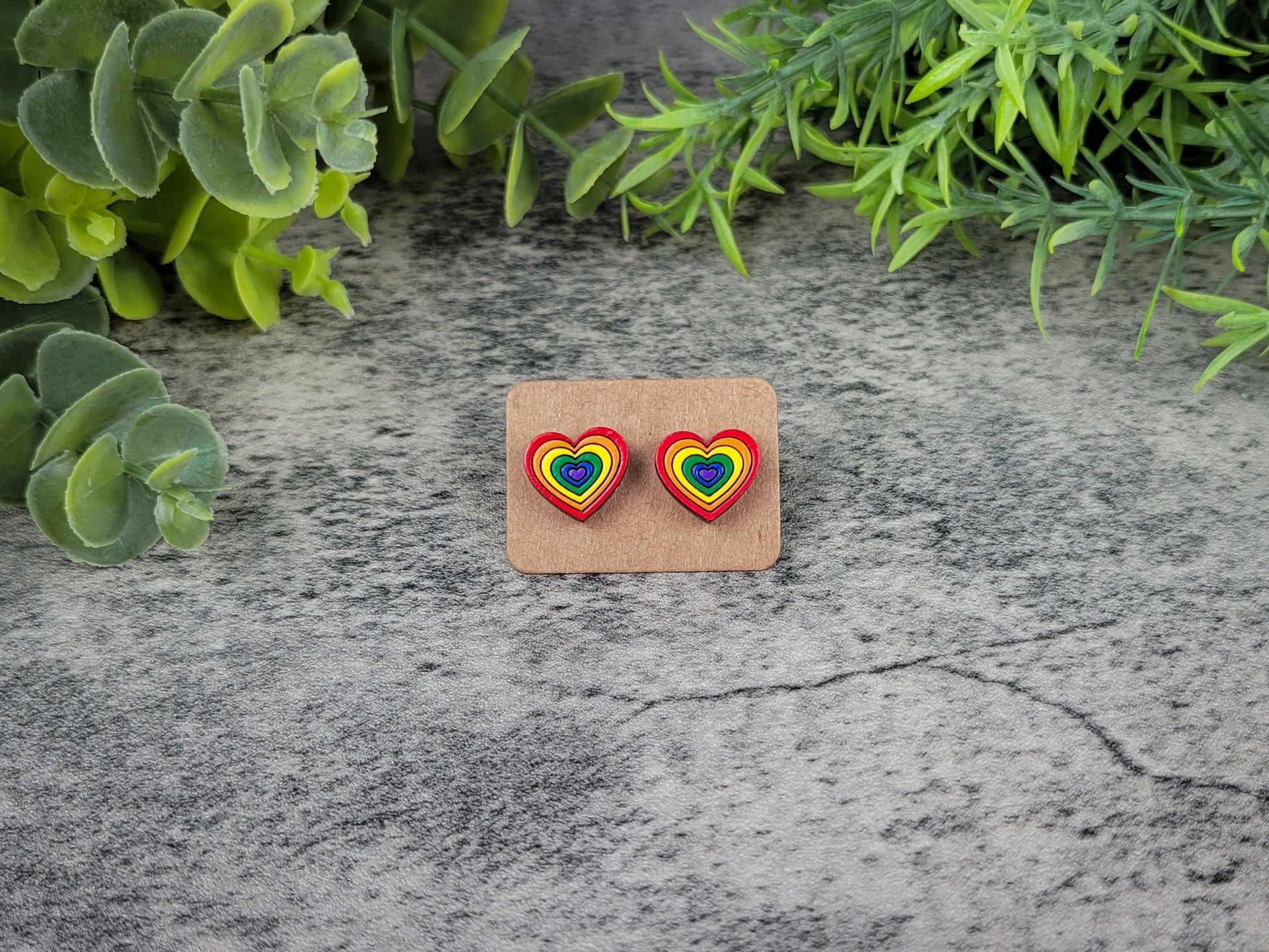 Heart-shaped earrings with rainbow design on a cardboard backing, placed on a textured stone surface with green foliage.