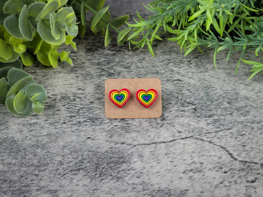 Heart-shaped earrings with rainbow design on a cardboard backing, placed on a textured stone surface with green foliage.