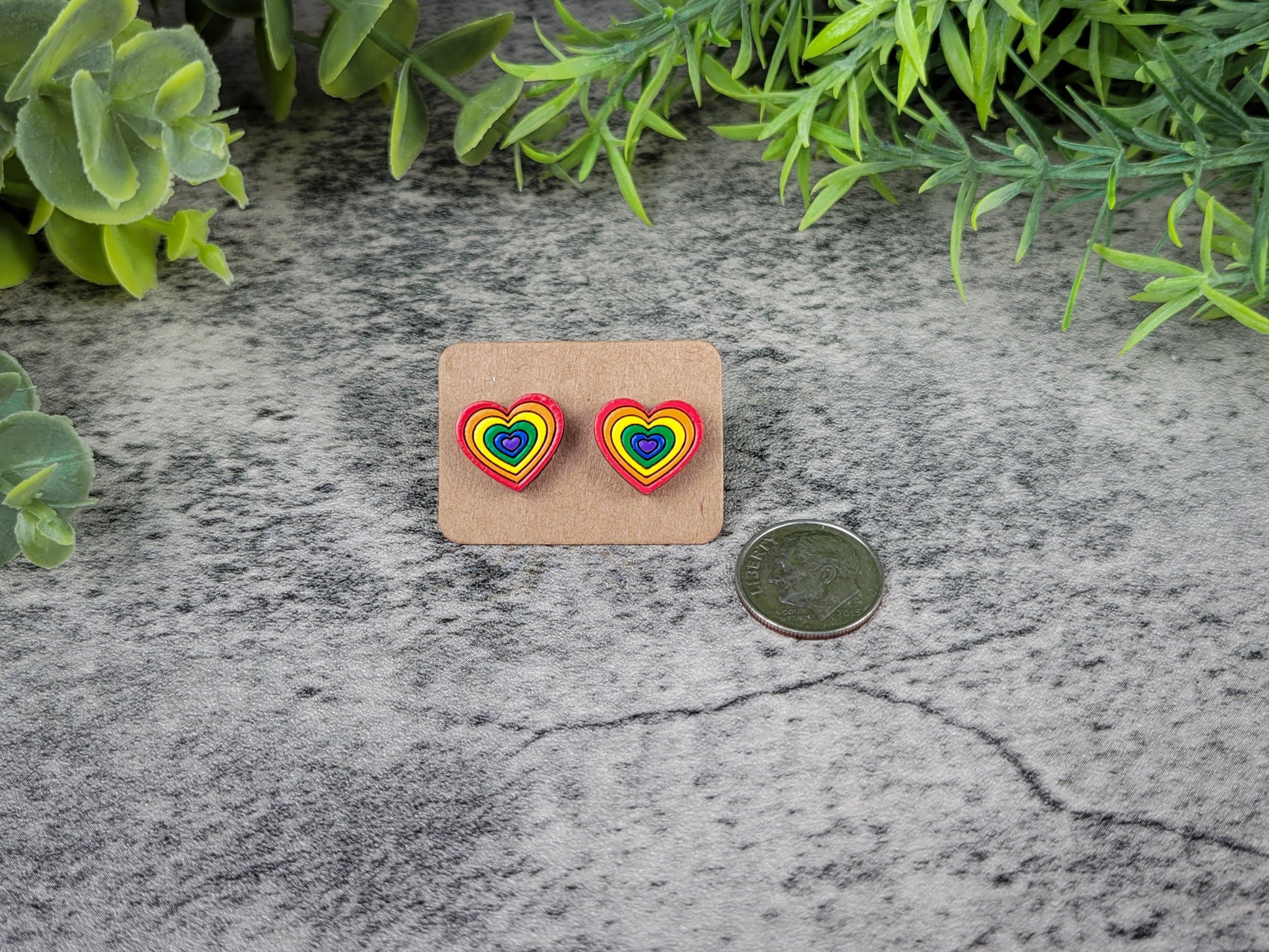 Rainbow heart-shaped earrings on a cardboard backing with a coin for scale, placed on a textured surface with greenery.