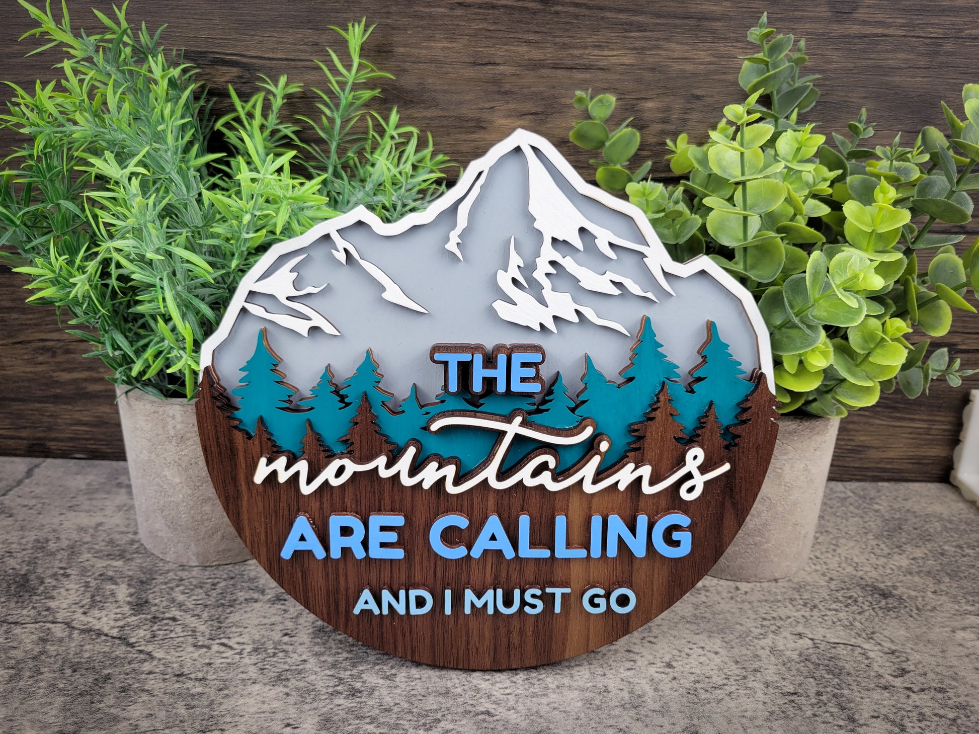 Decorative sign with mountain design and text 'The mountains are calling and I must go' surrounded by plants.