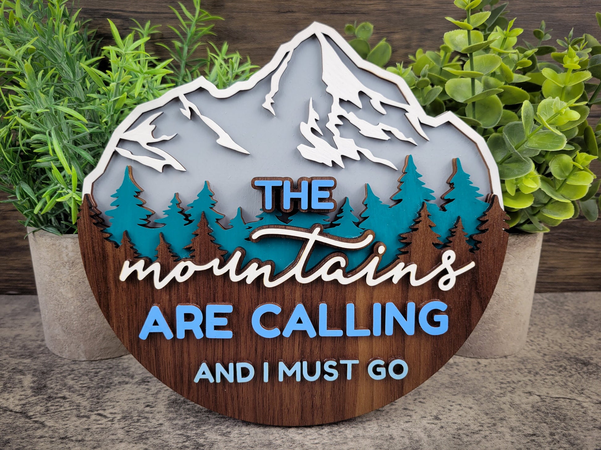 Decorative sign with mountain design and text 'The mountains are calling and I must go' surrounded by plants.
