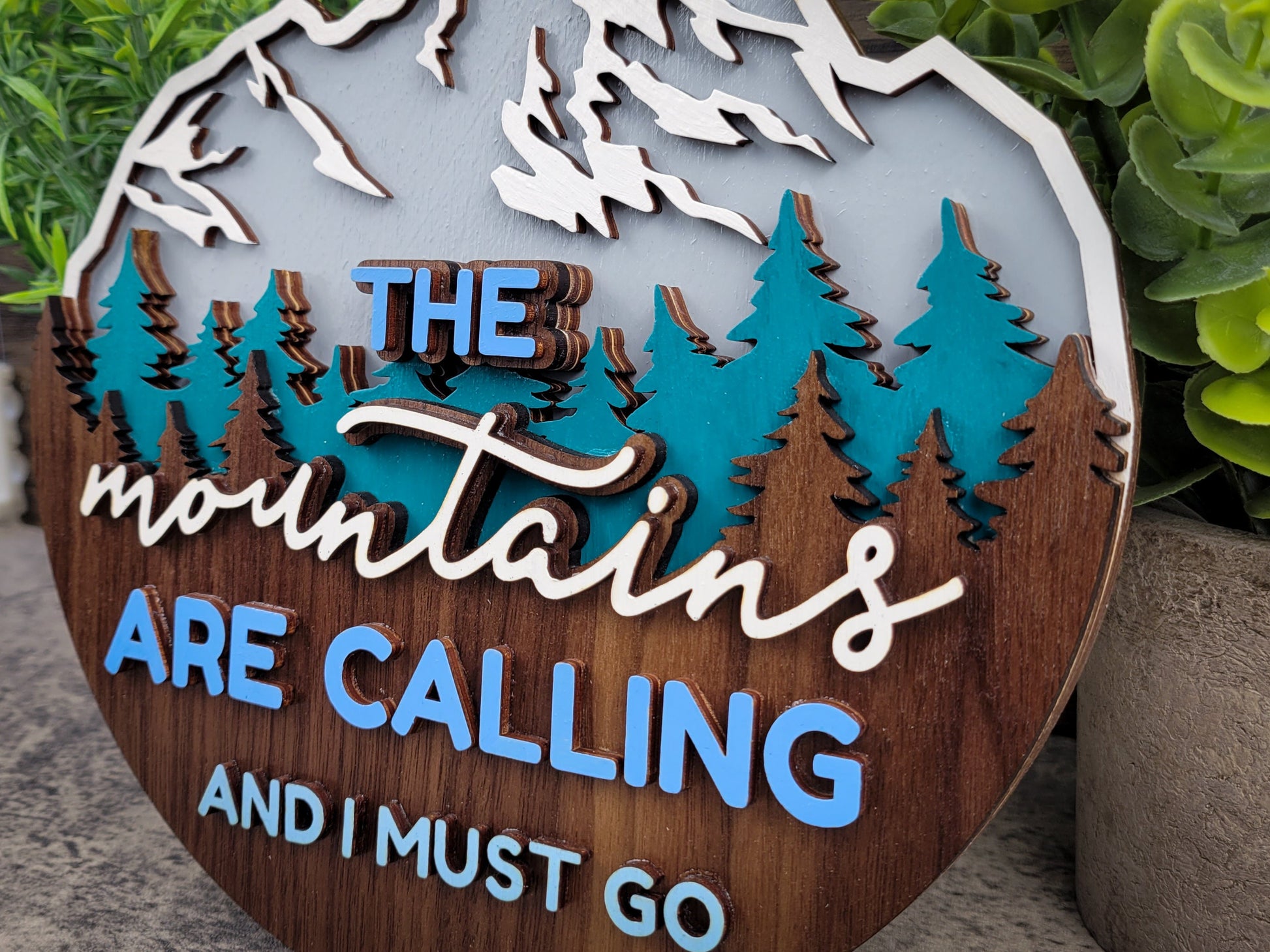 Decorative sign with 'The mountains are calling and I must go' text surrounded by trees.