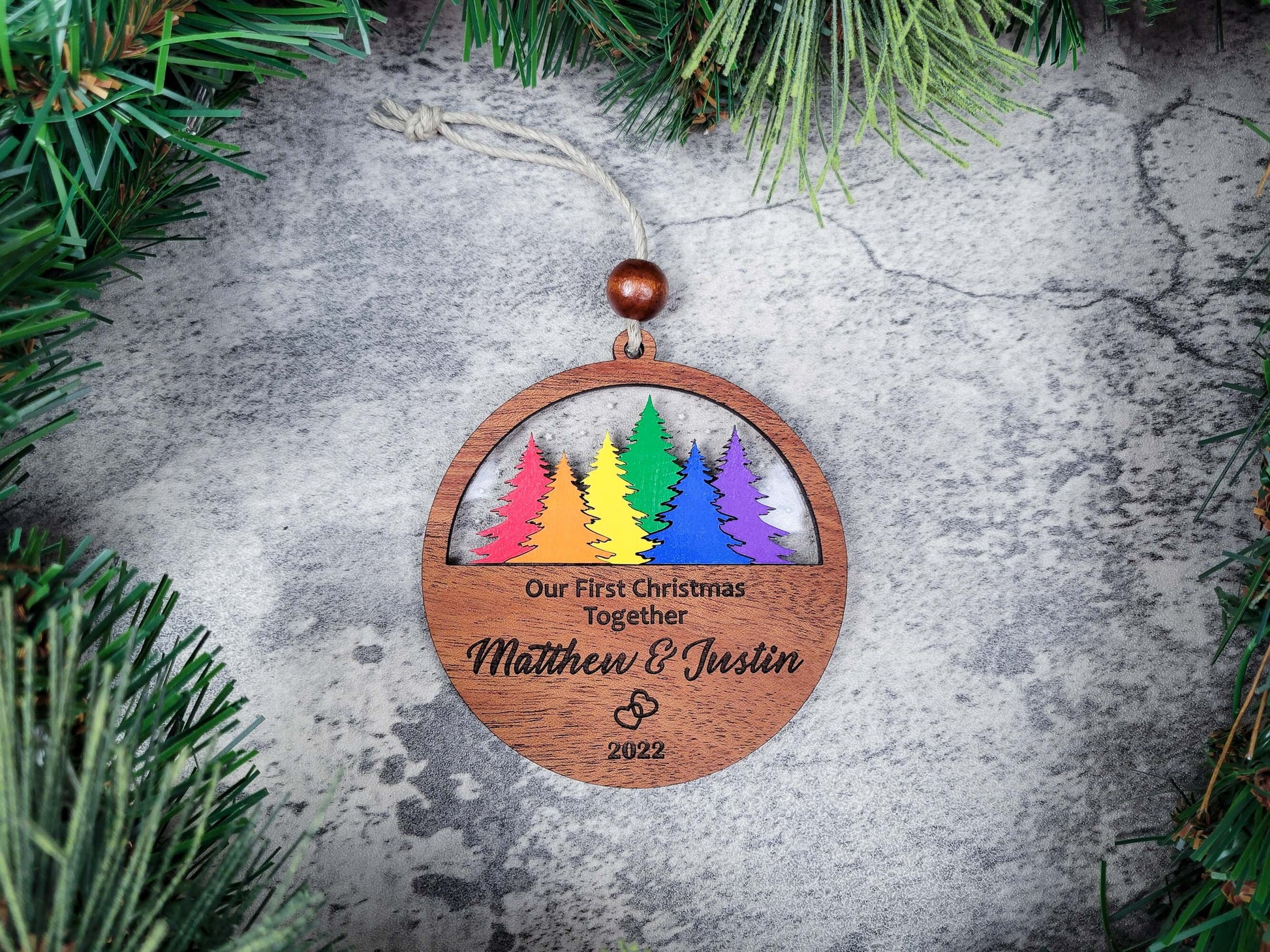 Wooden Christmas ornament with colorful trees and personalized text on a stone surface with greenery.