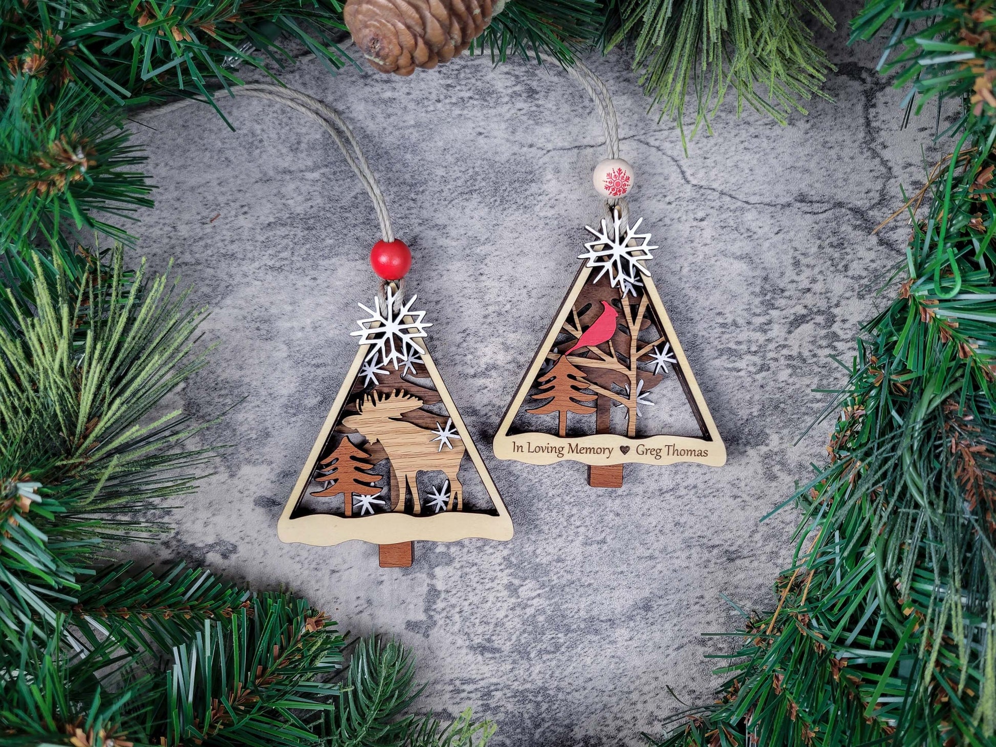 Two wooden Christmas tree ornaments with engraved designs on a textured surface surrounded by pine branches.
