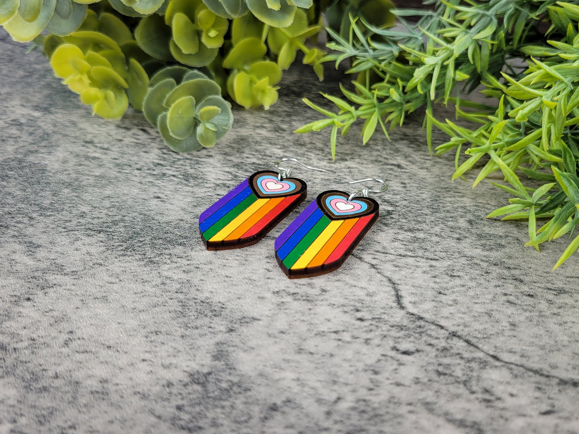 Rainbow heart-shaped earrings on a textured surface with green foliage in the background