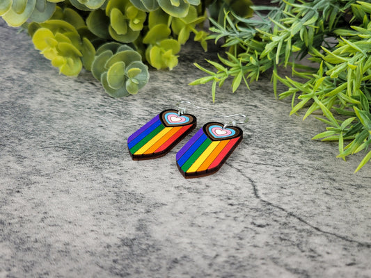 Rainbow heart-shaped earrings on a textured surface with green foliage in the background