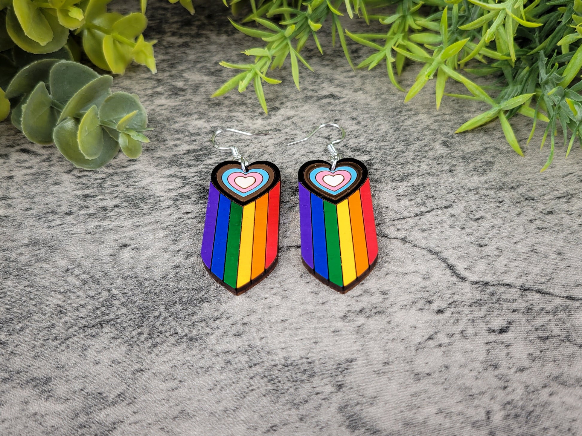 Rainbow heart-shaped earrings on a textured surface with greenery in the background