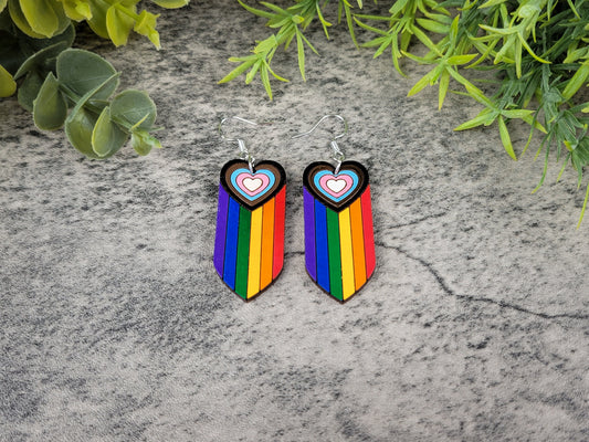 Rainbow heart-shaped earrings on a textured surface with green leaves.