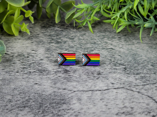 Pair of rainbow flag earrings on a textured surface with greenery in the background