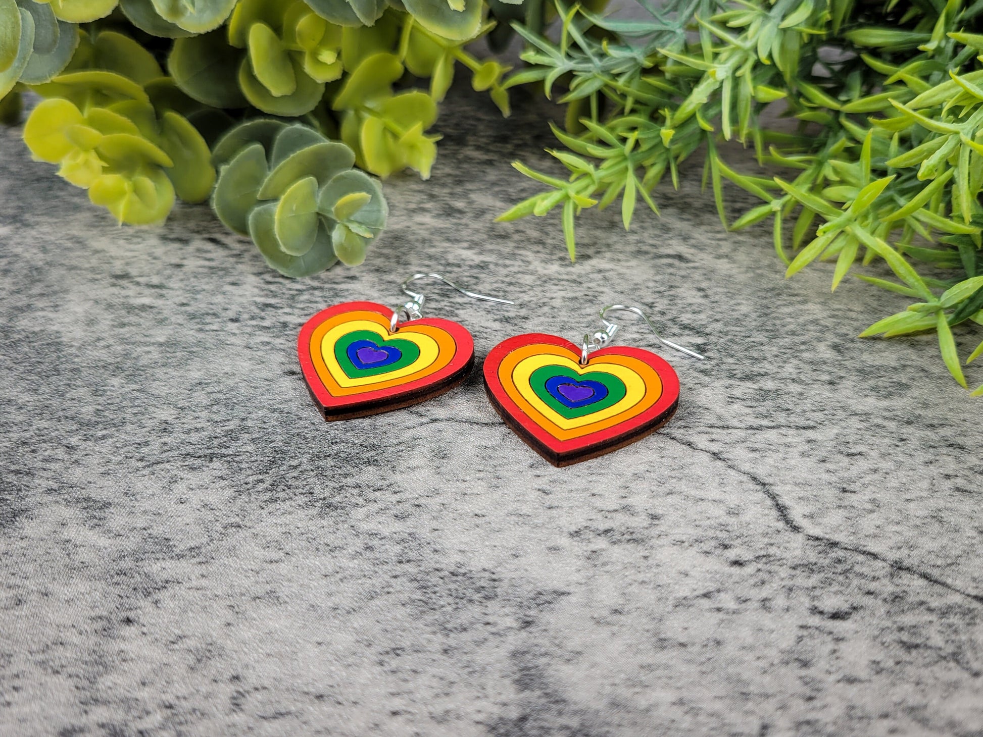 Rainbow heart-shaped earrings on a stone surface with green foliage in the background
