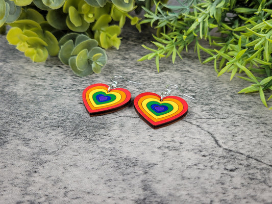 Rainbow heart-shaped earrings on a stone surface with green foliage in the background