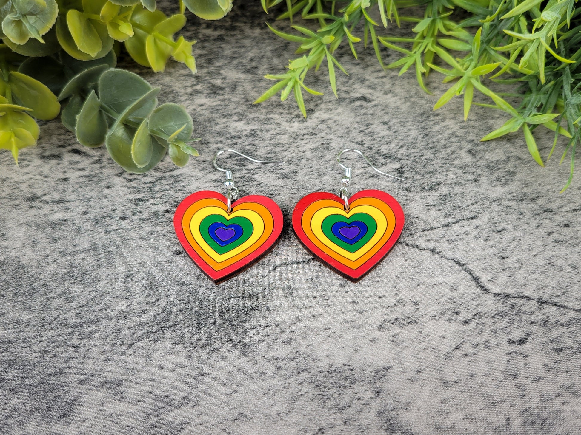 Rainbow heart-shaped earrings on a textured surface with greenery