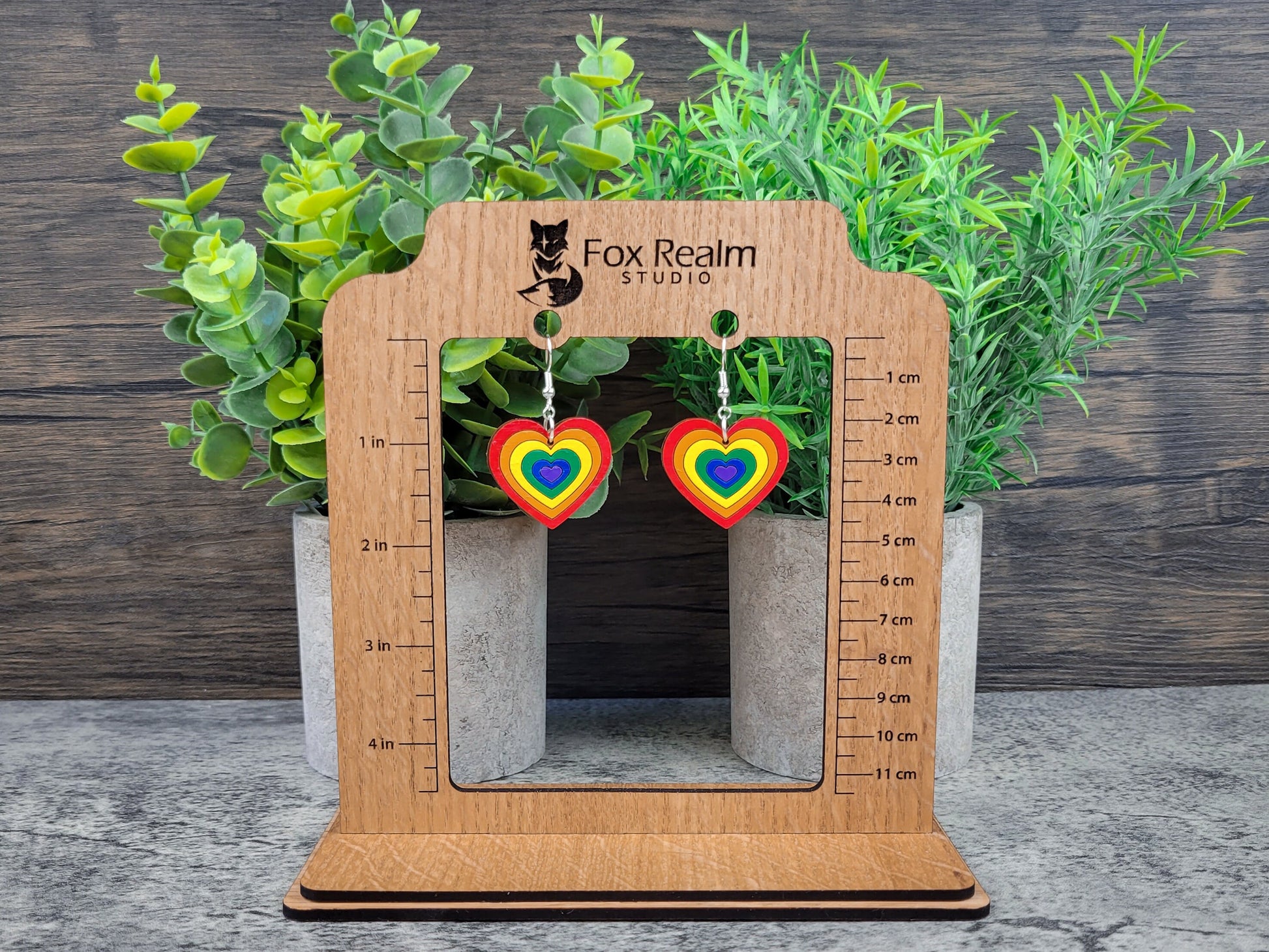 Heart-shaped rainbow earrings on a wooden measurement stand with plants in the background