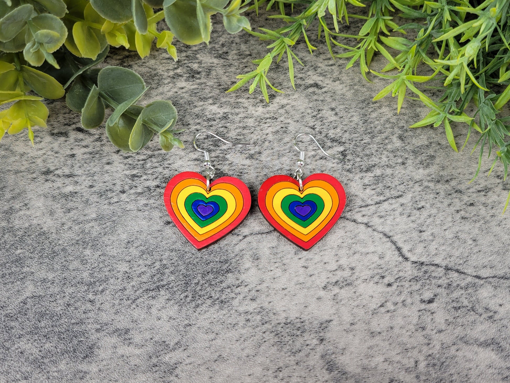 Rainbow heart-shaped earrings on a textured stone surface with green foliage.