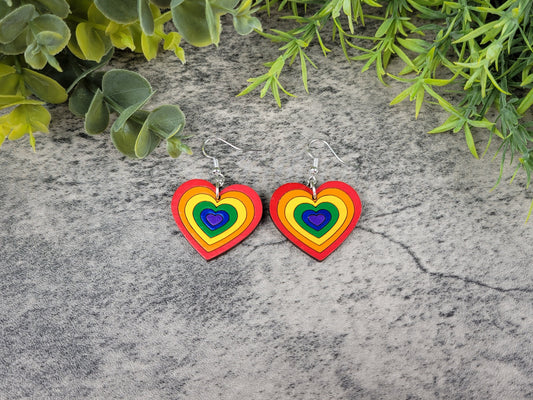Rainbow heart-shaped earrings on a textured stone surface with green foliage.