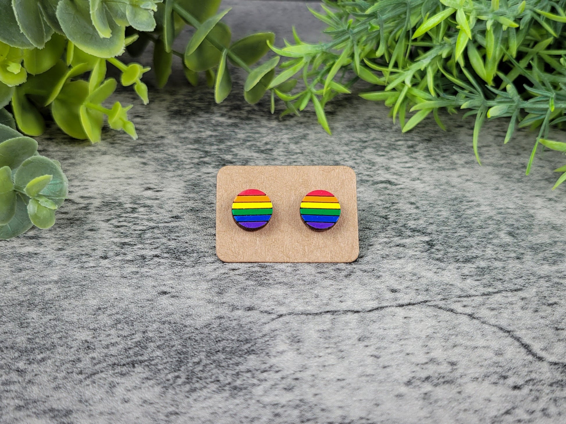 Rainbow-colored earrings on a cardboard backing with green leaves in the background