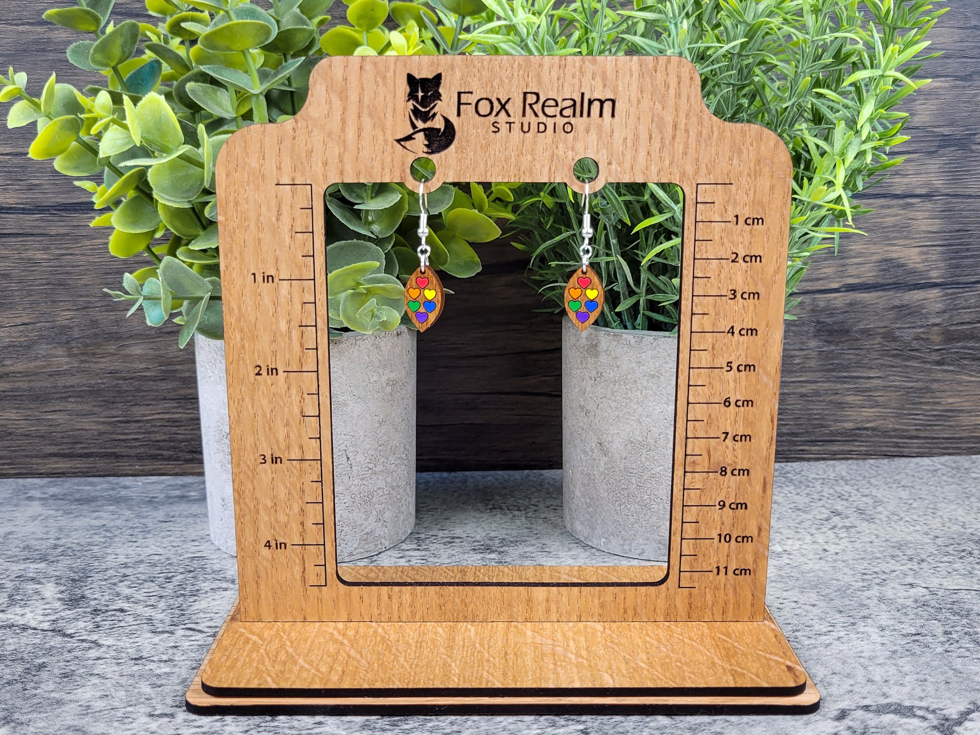 Wooden earring display stand with measurement scale, featuring Fox Realm Studio branding, against a wooden background.