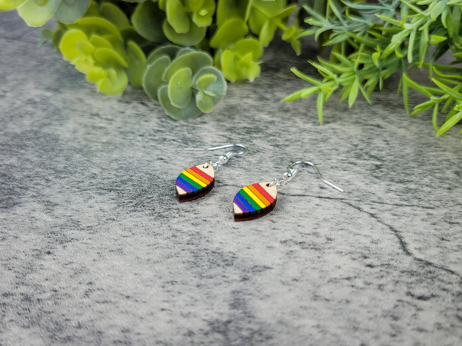 Rainbow heart-shaped earrings on a gray surface with greenery in the background