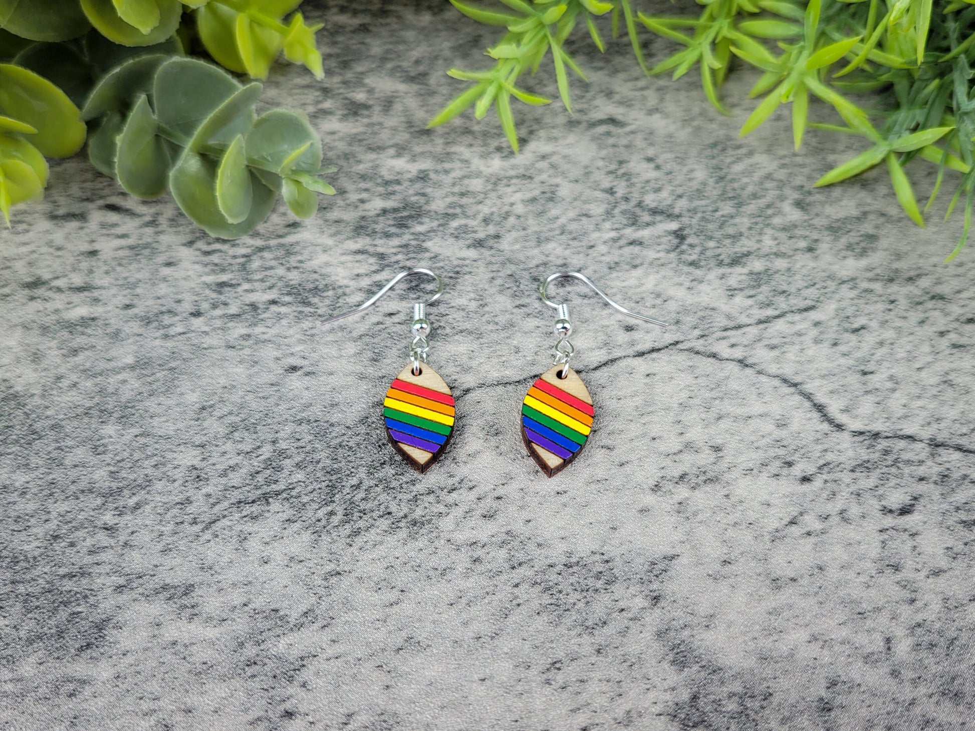 Rainbow fish-shaped earrings on a textured stone surface with greenery.