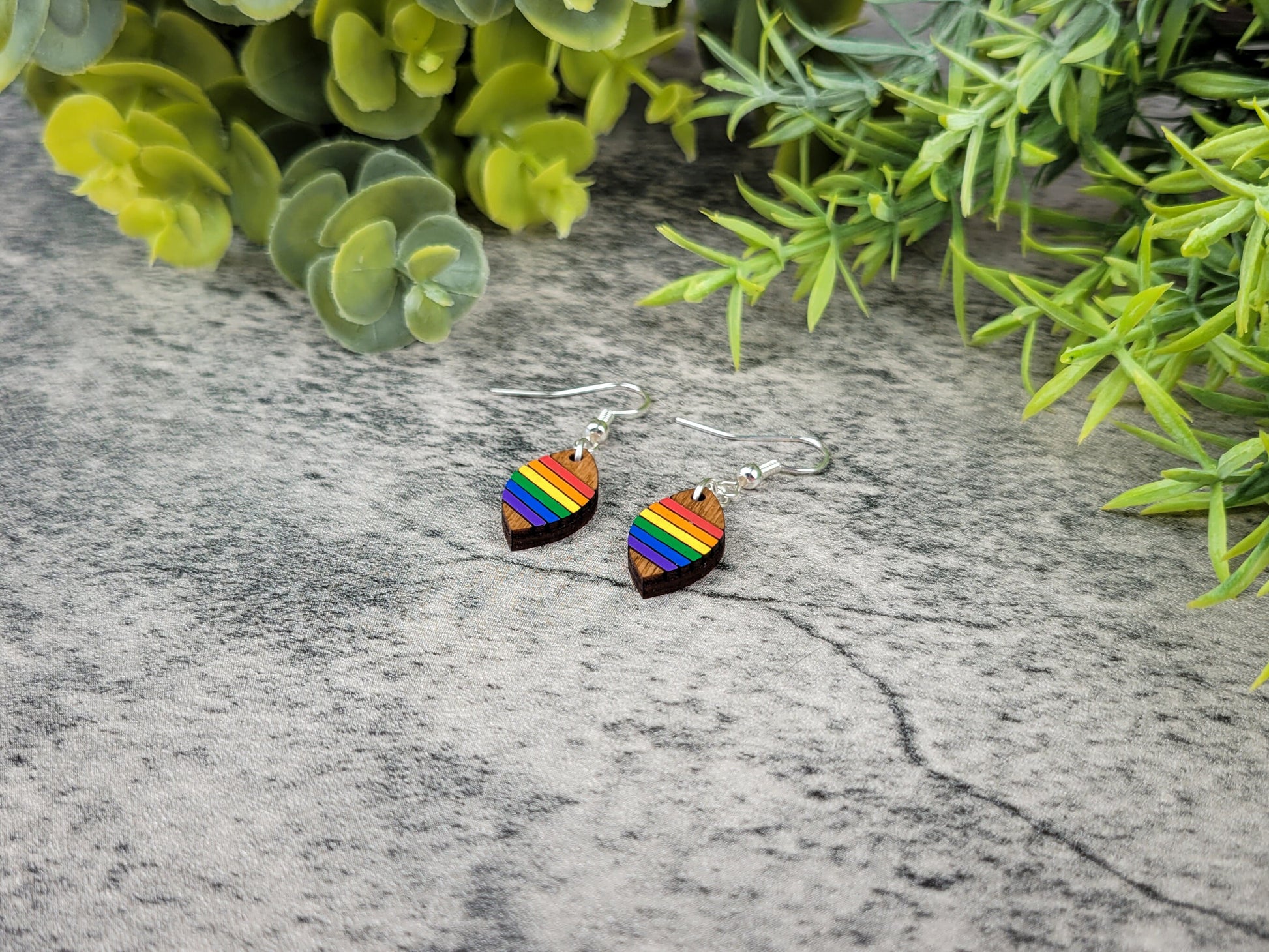 Rainbow heart-shaped earrings on a textured surface with green foliage in the background
