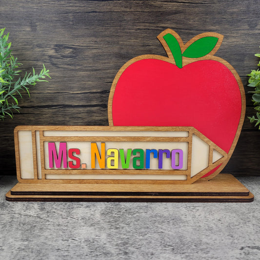 Wooden nameplate with colorful letters spelling 'Ms. Navarro' and a red apple on a wooden background.