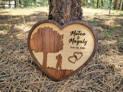 Wooden heart-shaped plaque with engraved tree and hearts, placed against a tree trunk in a forest setting.