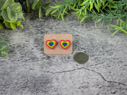 Rainbow heart-shaped earrings on a cardboard backing with a coin for scale, placed on a textured surface with greenery.