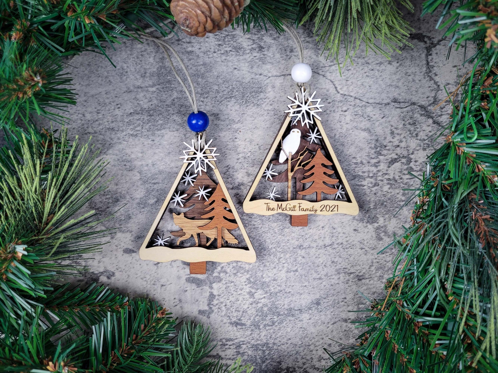 Two triangular wooden Christmas tree ornaments with nature-themed designs on a textured surface surrounded by pine branches.