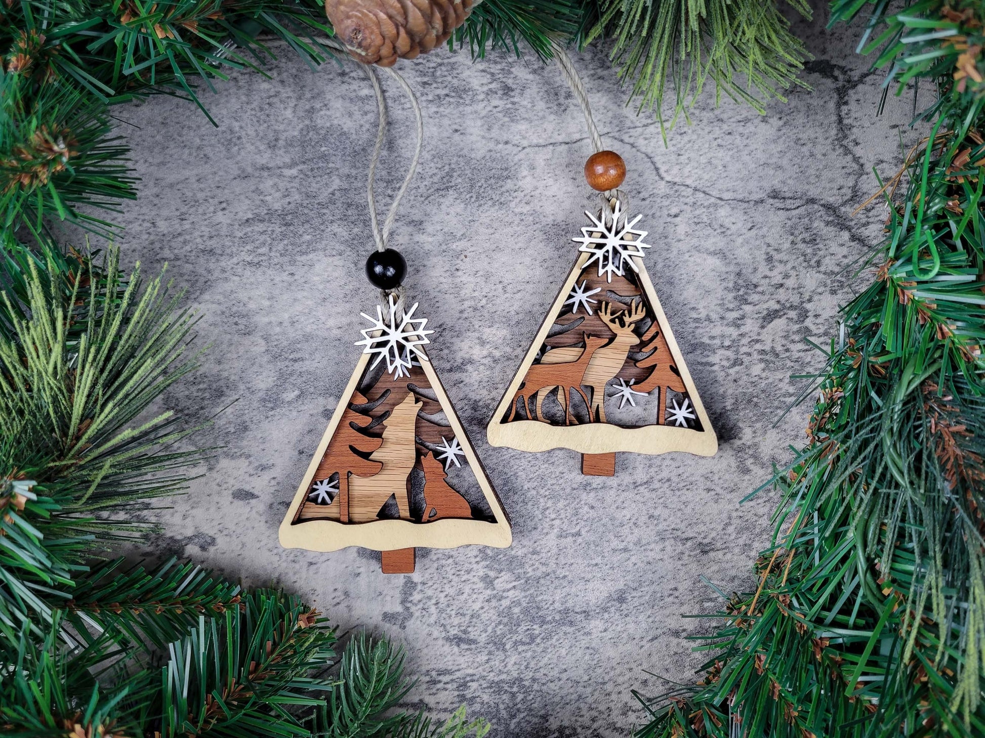 Two wooden Christmas tree ornaments with reindeer designs on a textured surface surrounded by pine branches.