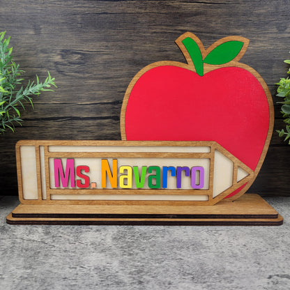 Wooden nameplate with colorful letters spelling 'Ms. Navarro' and a red apple on a wooden background.